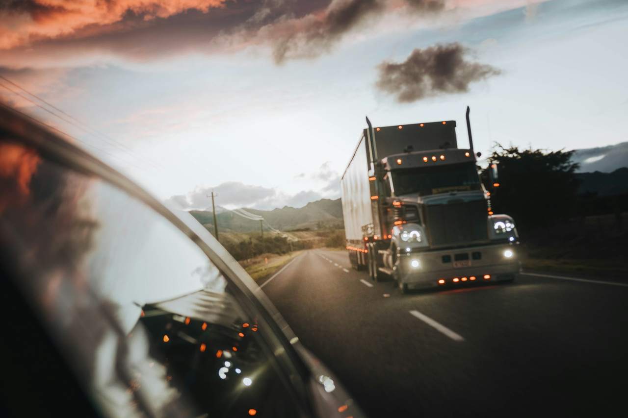 A semi truck with its headlights on, driving on a highway at dusk, seen in a side mirror.
